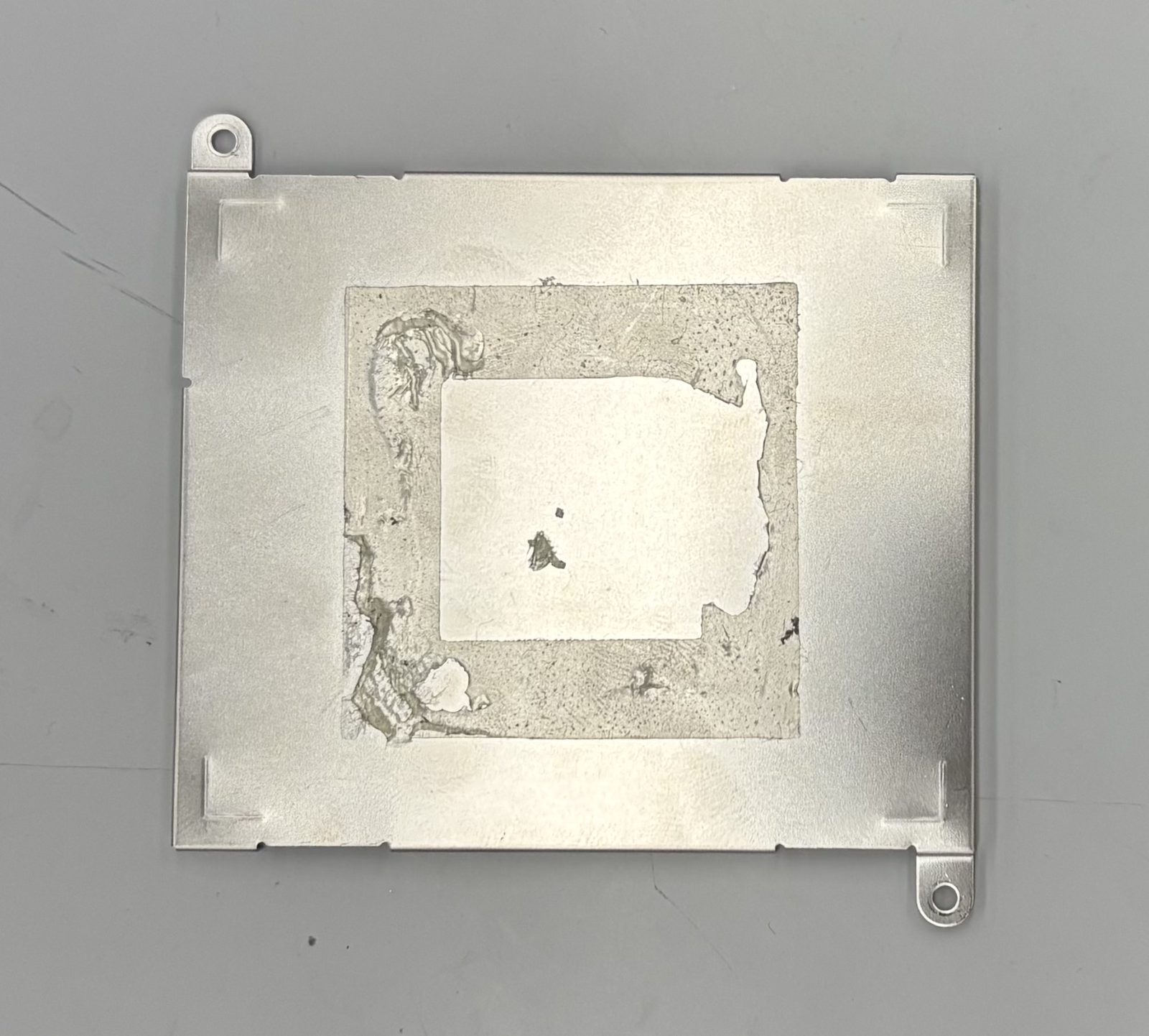 Bare metal side of the battery cover with adhesive residue in the centre and screw tabs on two corners