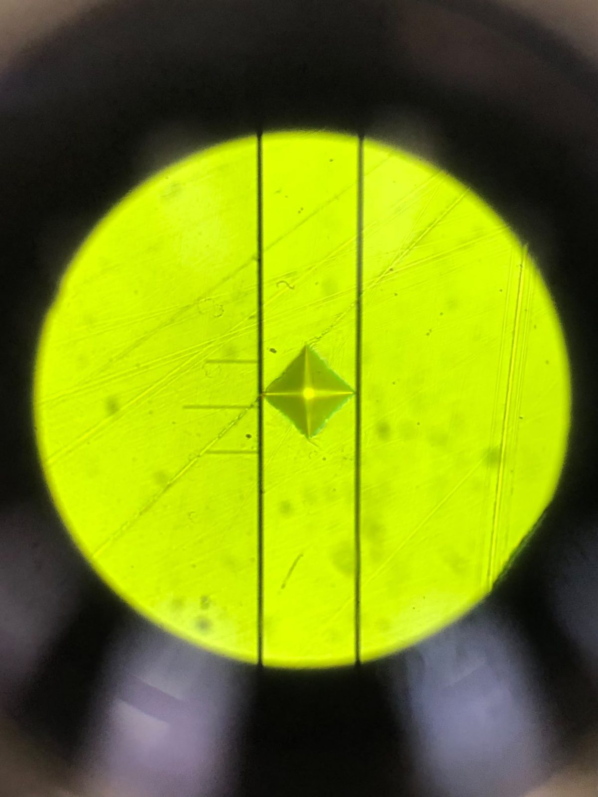 Microscope view through the eyepiece of a diamond-shaped Vickers indent between two measurement lines