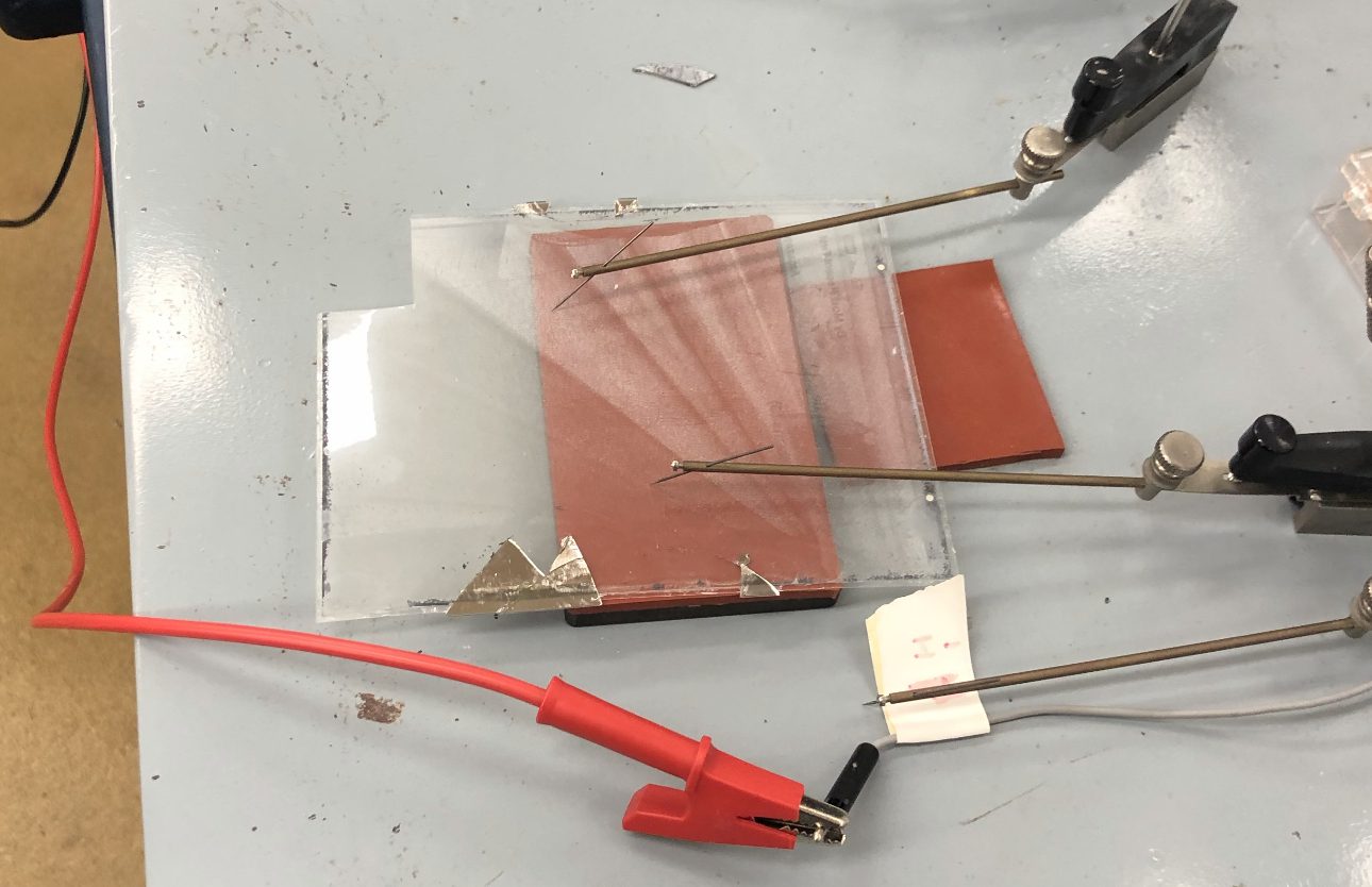 Two probe needles resting on the clear film, connected to a power supply by red leads